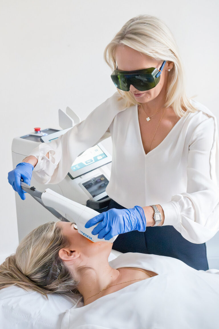 A woman wearing protective goggles and blue gloves uses a handheld laser device on another woman's face, who is lying down with her eyes closed, during a cosmetic treatment in a medical setting.