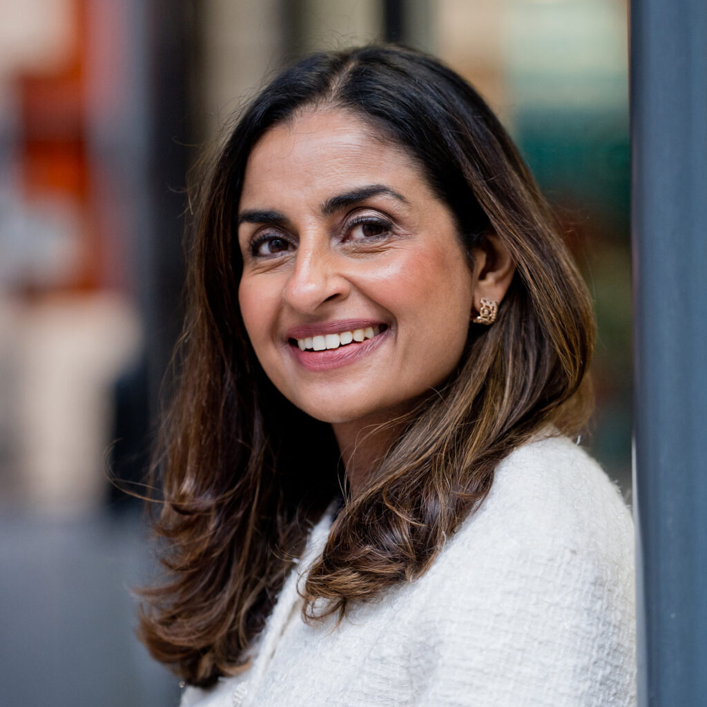 A woman with long brown hair, wearing a white textured jacket and gold earrings, smiles whilst standing outdoors near a metal pole. The background is softly blurred.