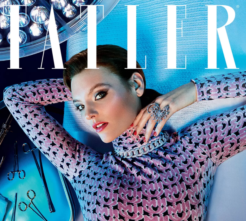 A woman in a patterned pink and purple outfit reclines on a blue surface, gazing up with her hand near her chin, wearing bold make-up and statement rings. Large white TATLER text is overlaid at the top.