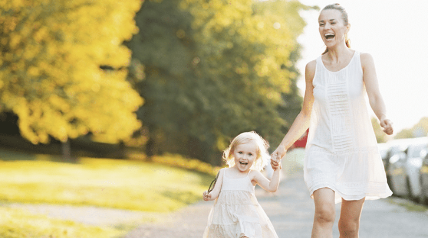 A woman and a young girl, both in light-coloured dresses, are holding hands and smiling whilst walking outdoors on a sunny day with green trees and parked cars in the background.