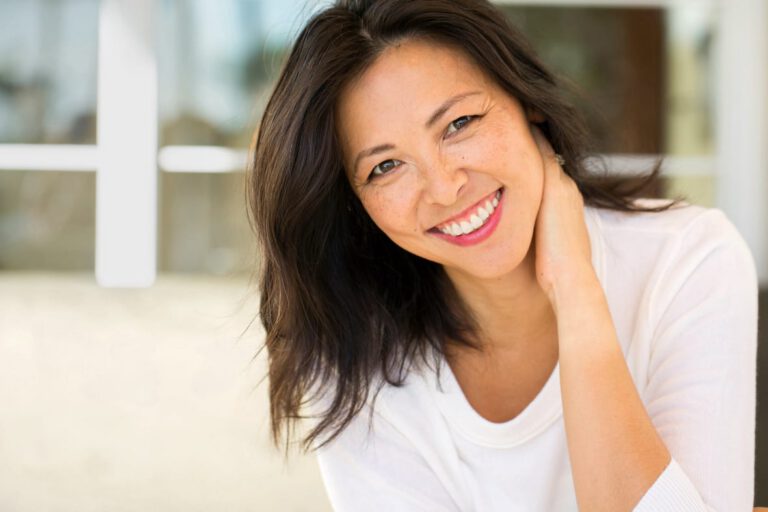 A woman with long dark hair and a white top smiles warmly at the camera, tilting her head and resting one hand on her neck. The background is softly blurred, suggesting an indoor setting with natural light.