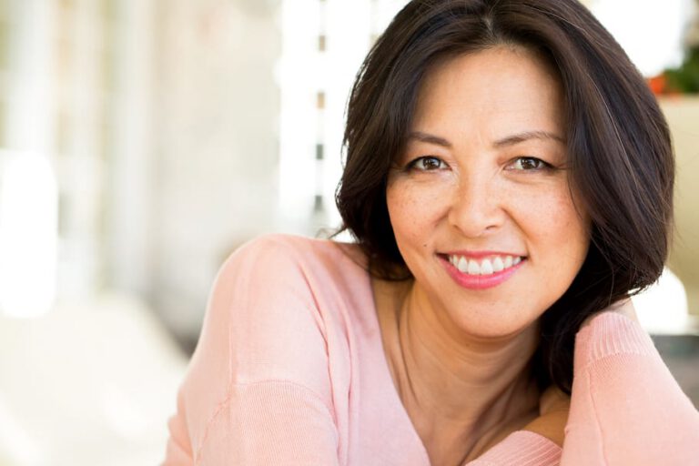 A woman with long dark hair, wearing a light pink top, smiles warmly at the camera. She is indoors with a softly blurred background and her arm rests casually, supporting her head.