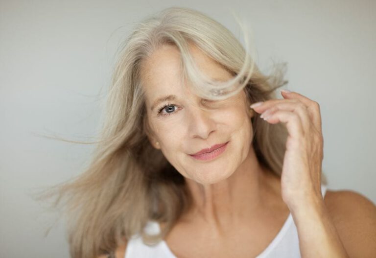 A smiling older woman with long, grey hair wears a white top. Some hair is gently blowing across her face as she lifts her hand, creating a soft, natural look against a plain background.