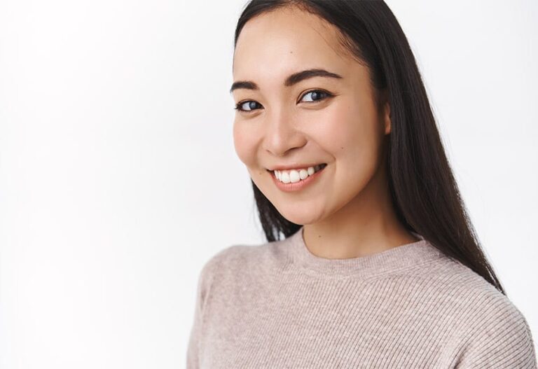 A young woman with long dark hair smiles at the camera. She is wearing a light pink ribbed jumper and stands against a plain white background.