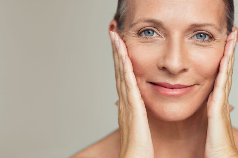 Close-up of a middle-aged woman with light skin smiling gently, touching both sides of her face with her hands against a neutral background. Her expression is calm and contented.