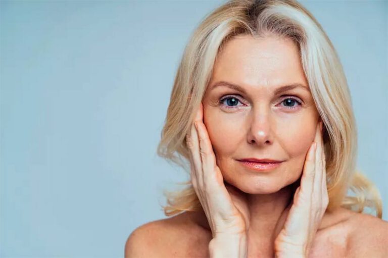 A middle-aged woman with blonde hair touches both sides of her face gently, looking directly at the camera, against a plain light blue background.
