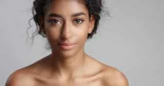 A young woman with curly dark hair and clear skin looks directly at the camera, smiling softly. She has bare shoulders and is posed against a plain, light grey background.