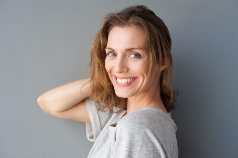 Smiling woman with wavy brown hair wearing a grey t-shirt stands against a plain grey wall, looking over her shoulder at the camera with a relaxed and cheerful expression.