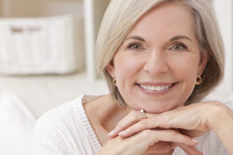 Smiling older woman with short grey hair rests her chin on her hands. She wears a white jumper and gold hoop earrings, with a ring visible on her finger. The background is softly blurred.