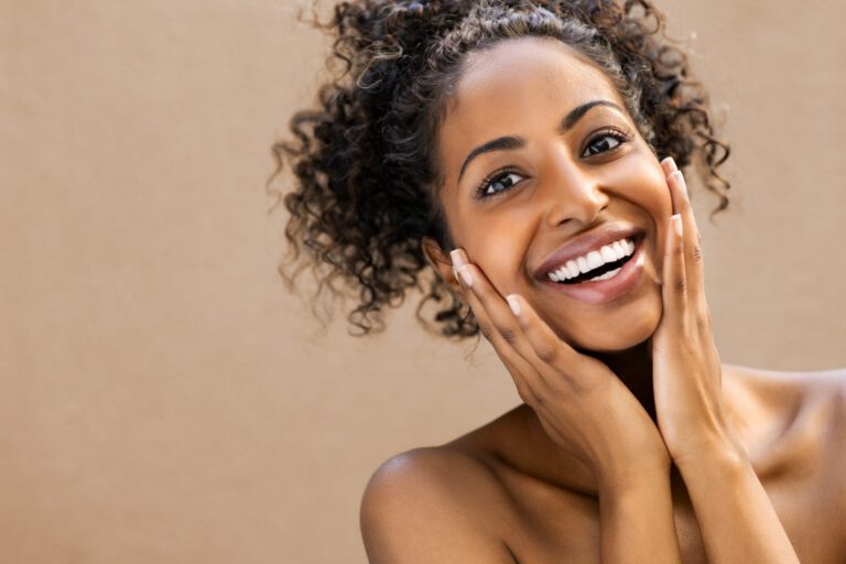 A young woman with curly hair smiles brightly, touching her face with both hands. She has glowing skin and is posed against a plain, tan background.
