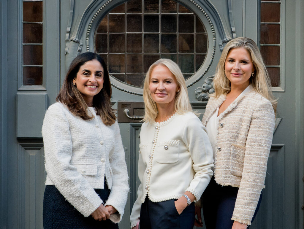 Three women in light-coloured jackets stand smiling in front of an ornate wooden door with a round window. They are positioned close together, facing the camera, and appear to be professional and confident.