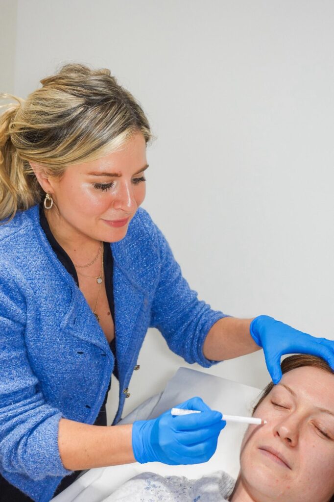 A blonde female aesthetician wearing blue gloves and a blue jacket uses a white marking pen to map a reclining patient's face before a cosmetic treatment.