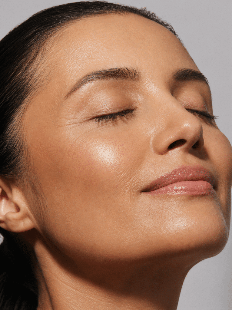 A close-up of a woman with smooth, glowing skin and closed eyes, smiling softly. Her head is slightly tilted back, and she appears calm and relaxed against a neutral background.